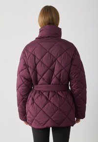 Giacca imbottita trapuntata prugna con colletto alto, vita con cintura e motivo a diamante; texture liscia e design di lunghezza intermedia.