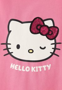 T-shirt rosa con una grafica di un volto di gatto bianco che fa l'occhiolino, baffi neri e un fiocco rosa testurizzato, con "HELLO KITTY" in testo bianco sottostante.