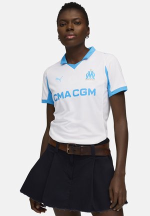 Vit fotbollsdräkt med ljusblå detaljer och krage, med "CMA CGM" i fet blå text och en logotyp. Matchas med en svart kjol.