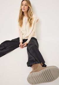 Gilet en polaire crème avec fermeture éclair à l'avant, superposé à un top à manches longues beige clair. Jean noir à jambes larges et bottines tan avec semelles texturées.