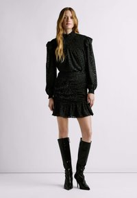 Vestido negro texturizado con mangas abullonadas, cintura fruncida y dobladillo con volantes. Combinado con botas negras brillantes hasta la rodilla.