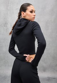 Joven con cabello largo trenzado, vestida con un crop top de capucha negro y pantalones, girando para mirar por encima del hombro contra una pared gris.