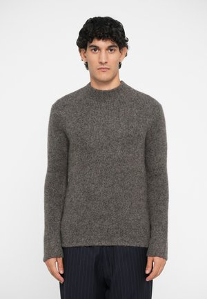 Brauner Strickpullover mit Rundhalsausschnitt, langen Ärmeln und strukturiertem Stoff, kombiniert mit dunkel gestreiften Hosen.