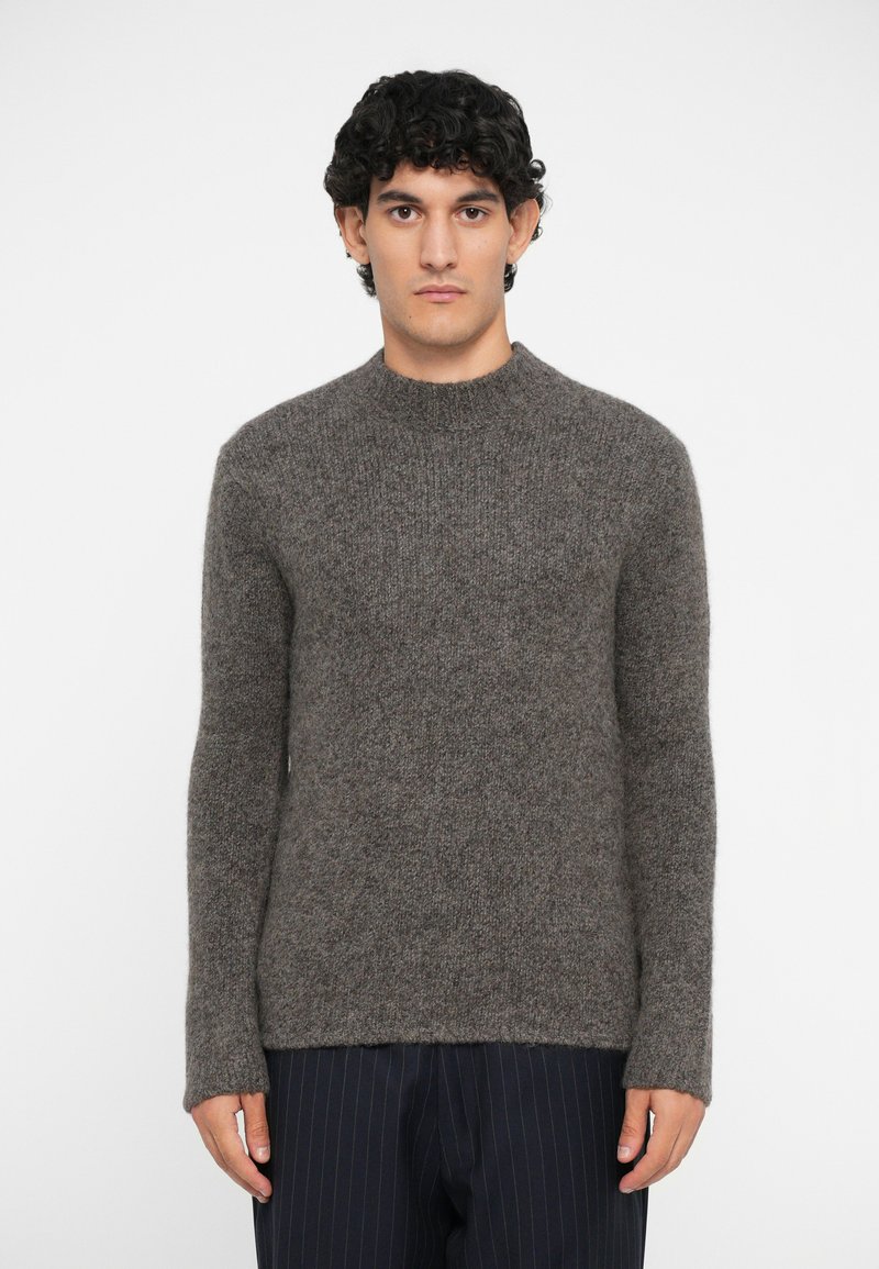 Brown knit sweater with a crew neck, long sleeves, and textured fabric, paired with dark pinstriped trousers.