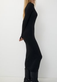 Femme dans une robe noire ajustée à manches longues et des bottes noires se tient de profil contre un fond blanc uni.