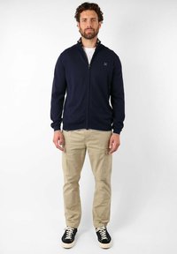 Veste zippée bleu marine avec des manches longues, en tissu texturé et un petit logo. Associée avec un pantalon cargo beige et des baskets noires.