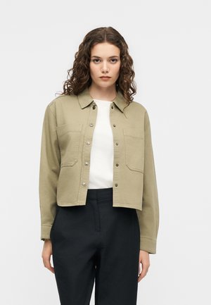 Jeune femme aux cheveux bouclés portant une veste beige boutonnée sur un haut blanc et un pantalon noir, debout devant un fond uni.