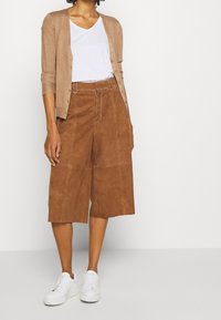 Bruine suède culotte broek met voorzakken en witte stiksels, gecombineerd met een beige vest over een wit V-hals T-shirt en witte sneakers.