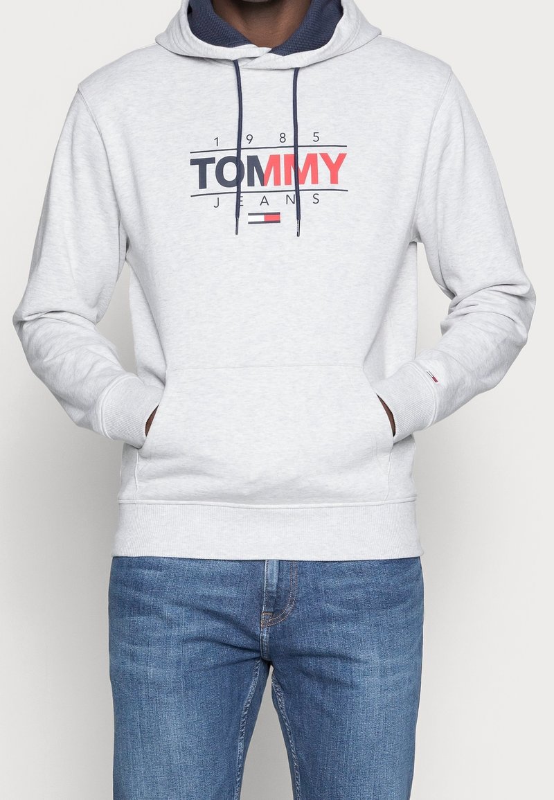 Sudadera gris claro con el logo "Tommy Jeans 1985" en navy y rojo, forro de la capucha en navy, cordones, bolsillo tipo canguro, combinada con jeans de mezclilla azules.