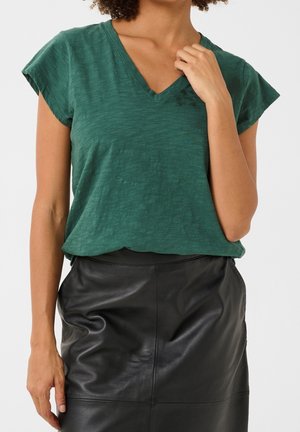Kobieta ubrana w zielony t-shirt z krótkim rękawem i dekoltem w serek, wpuszczony w wysoką, czarną skórzaną spódnicę na jednolitym tle.