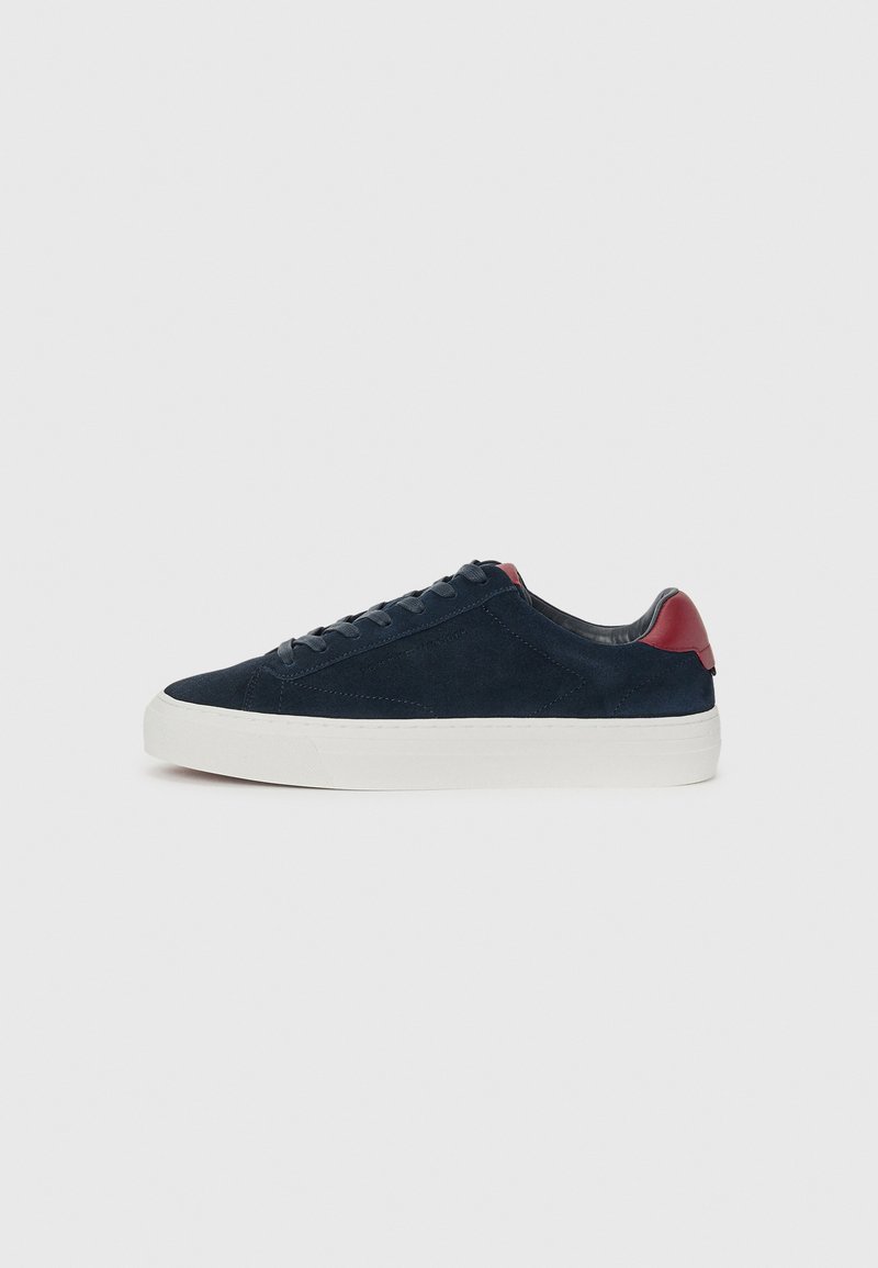 Marineblå sneakers i semsket skinn med hvit gummisåle, med rød hælkant og flate snørebånd, glatt tekstur og minimalistisk design.