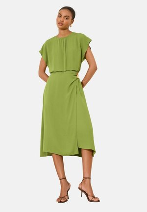 Kvinde iført en grøn midi-kjole med korte ærmer og en rynket talje, parret med brune hæle med stropper.