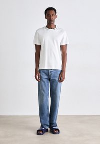 T-shirt blanc à manches courtes avec un col bordé de vert, associée à un jean droit bleu clair et des sandales à glissière bleu marine avec un texte blanc.
