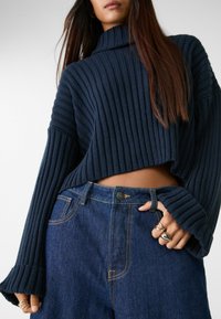 Mornaricki rebrasti pletivi turtleneck pulover s širokim rukavima, uparen s visokim plavim trapericama koje sadrže zlatne šavove i metalne detalje.