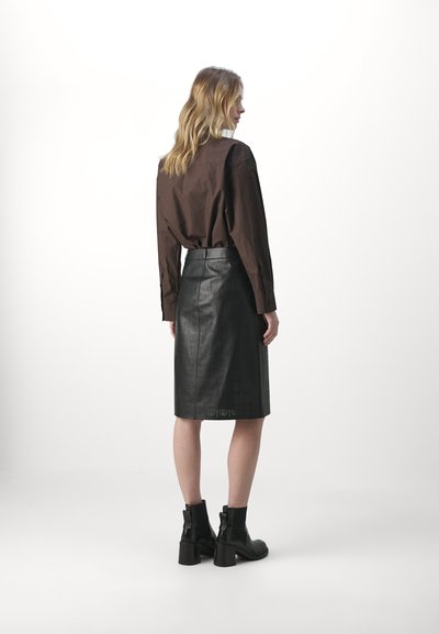Zwarte leren kokerrok met ingegraveerde patronen, hoge taille en een gladde textuur. Combinatie met een bruine blouse met kraag en zwarte enkellaarsjes.