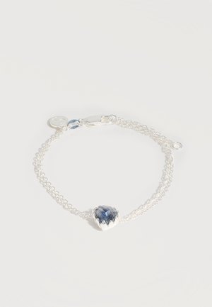 Λεπτό ασημένιο βραχιόλι με διπλή αλυσίδα, κεντρικό διακοσμητικό καρδιά με μπλε πολύτιμη πέτρα και μικρό στρογγυλό γούρι κοντά στο κούμπωμα.