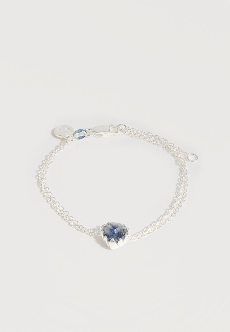 Λεπτό ασημένιο βραχιόλι με διπλή αλυσίδα, κεντρικό διακοσμητικό καρδιά με μπλε πολύτιμη πέτρα και μικρό στρογγυλό γούρι κοντά στο κούμπωμα.