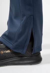 Marineblauwe sportbroeken met getextureerde zijkanten en ritsen bij de enkels, gecombineerd met zwarte sportschoenen met een patroon.