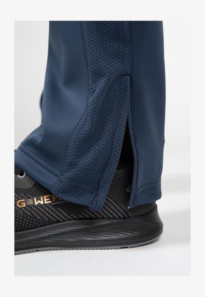 Marineblauwe sportbroeken met getextureerde zijkanten en ritsen bij de enkels, gecombineerd met zwarte sportschoenen met een patroon.