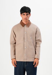 Man wearing beige buttoned jacket with brown collar and front pockets, paired with dark blue jeans, standing against a white background.