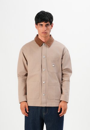 Man wearing beige buttoned jacket with brown collar and front pockets, paired with dark blue jeans, standing against a white background.
