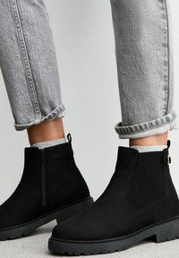 Svarta mocka ankboots med elastiska sidopaneler, texturerad gummisula och bakre dragband, som bärs med rullade grå jeans och ljusa strumpor.