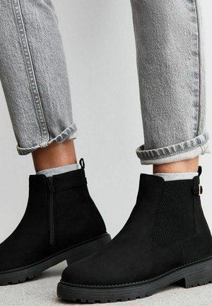 Bottes noires en daim à chevilles avec panneaux élastiques sur les côtés, semelle en caoutchouc texturée et languette à l'arrière, associées à un jean gris retroussé et des chaussettes claires.