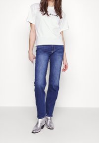 Witte katoenen t-shirt met "Dalkin" bedrukt, gecombineerd met blauwe spijkerbroek en zilverkleurige metallic enkelboots met lage hakken.