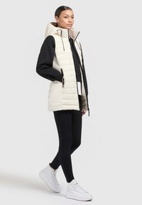 Gepolsterte, ärmellose Weste in hellem Creme mit Kapuze; kombiniert mit einem schwarzen Langarmoberteil und schwarzen Leggings; weiße High-Top-Schuhe.