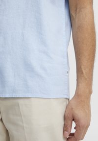 Camisa de manga corta azul claro en un tejido suave, con un dobladillo recto y una pequeña etiqueta en la costura lateral. Combinada con pantalones beige.