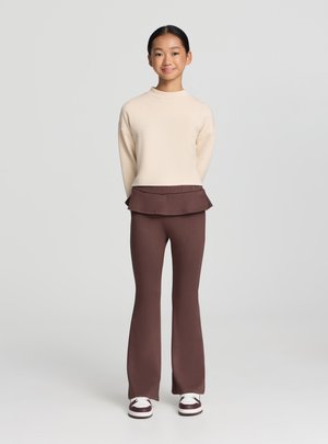 Maglietta crema a maniche lunghe con collo alto, vita a balza marrone stratificata; pantaloni aderenti marroni a zampetta; sneakers bianche con dettagli bordeaux.