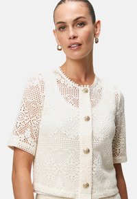 Cardigan bianco all'uncinetto con scollo rotondo, maniche corte e chiusure decorative a bottoni, caratterizzato da intricati motivi in pizzo e texture.