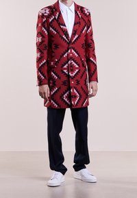 Long manteau rouge à motifs avec des designs géométriques en noir et blanc, associé à un pantalon foncé et des baskets blanches.