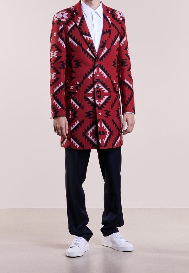 Long manteau rouge à motifs avec des designs géométriques en noir et blanc, associé à un pantalon foncé et des baskets blanches.