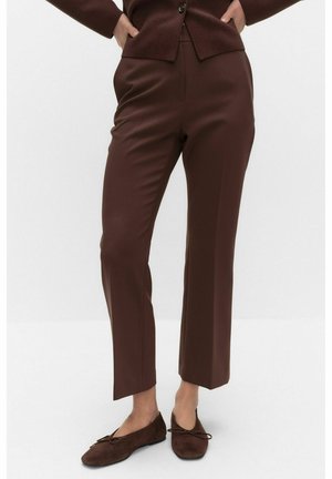 Femme portant un pantalon marron court sur mesure, un haut marron à manches longues et des ballerines en daim marron, debout les mains sur les hanches.
