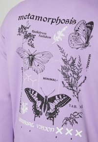 Maglietta viola con illustrazioni botaniche e di farfalle in bianco e nero, etichettata con nomi scientifici e parole come "metamorfosi" e "visione insolita."