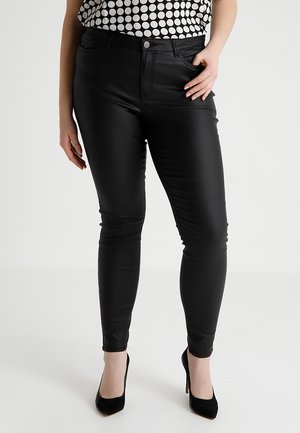 Femme portant un pantalon slim noir taille haute, des talons hauts noirs et un haut blanc sans manches à gros pois noirs.