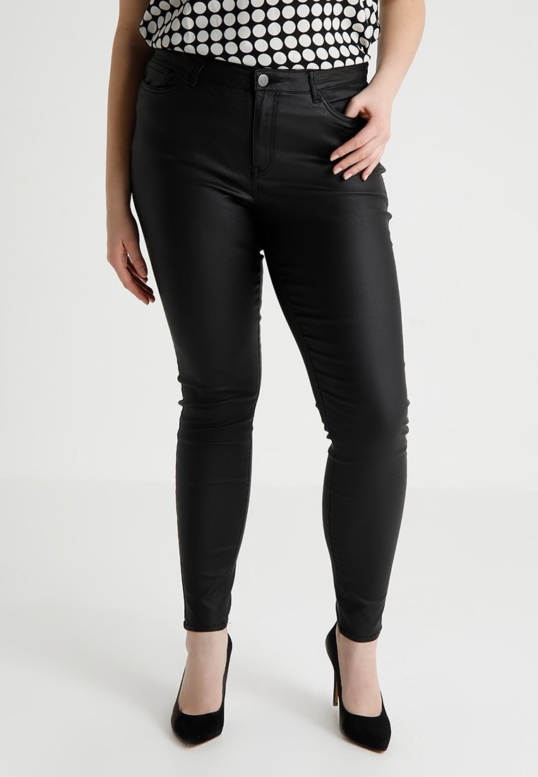 Femme portant un pantalon slim noir taille haute, des talons hauts noirs et un haut blanc sans manches à gros pois noirs.