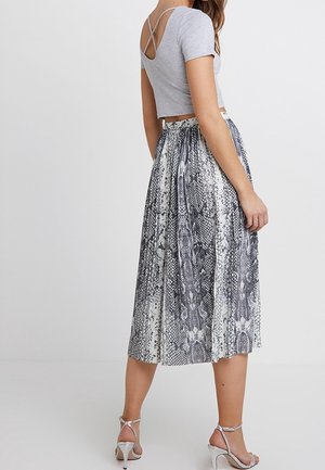 Kobieta ubrana w szary krótki crop top z krótkim rękawem, wzorzystą spódnicę midi i srebrne sandały na wysokim obcasie, stojąca na jednolitym tle.