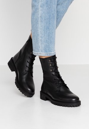 Personne portant un jean bleu clair et des bottes de combat en cuir noir à lacets, debout sur une surface blanche.