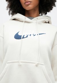 Sweatshirt com capuz creme com bolso frontal, apresentando um logótipo Nike azul texturizado em forma de 'swoosh' e o nome da marca bordado a branco. Detalhe de cordão ajustável no capuz.