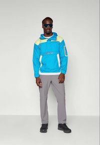 Blå og gul vindjakke med hætte, der har en frontlomme og sidekanter. Grå bukser og sorte sko fuldender outfittet.