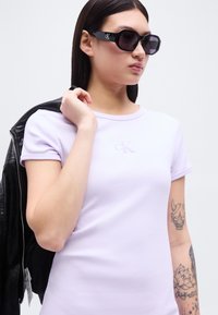 Νεαρός που φοράει μαύρα γυαλιά ηλίου Calvin Klein, ένα ανοιχτό μωβ t-shirt CK, κρατώντας ένα μαύρο σακάκι πάνω από τον έναν ώμο, με ορατά τατουάζ στο μπράτσο.