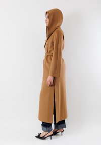 Lunga tunica marrone con cappuccio, cinturino, fessure laterali, maniche ampie e tasche, indossata sopra jeans scuri arrotolati e décolleté nere con cinturino.