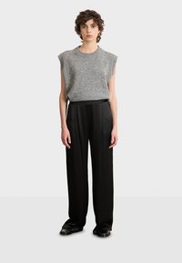 Grauer ärmelloser Strickoberteil mit geripptem Halsausschnitt, kombiniert mit schwarzen, hoch taillierten weiten Hosen. Das Modell steht mit den Händen seitlich.