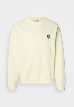 Sweatshirt amarelo claro com decote redondo, mangas compridas e um logótipo preto em forma de diamante no peito. Textura macia e corte confortável.