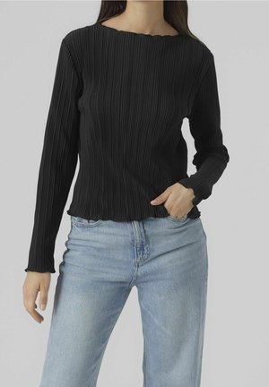 Vrouw draagt een zwart geribd shirt met lange mouwen en lichtblauwe jeans met hoge taille, één hand in de zak, neutrale achtergrond.