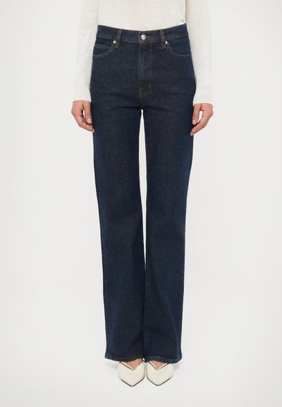 Jean denim bleu foncé à coupe décontractée, cinq poches et braguette zippée ; associé à des chaussures à bout pointu de couleur crème.