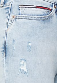 Vaqueros de mezclilla azul claro con parches desgastados, herrajes plateados y un pequeño detalle a rayas en rojo y marino en el bolsillo.