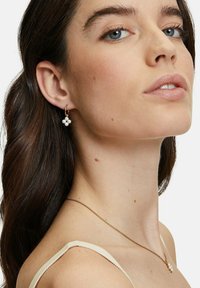 Junge Frau mit blauen Augen und dunklem Haar trägt goldene Perlenohrringe und eine passende Halskette vor einem weißen Hintergrund.